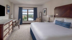Habitación de hotel con una cama grande, un escritorio, una silla, un televisor y vista a un cuerpo de agua y edificios.