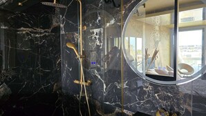 Un baño moderno con pared de mármol, cabezal de ducha dorado y un espejo redondo que refleja la habitación.