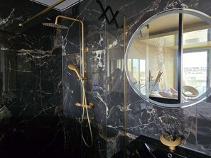 A modern bathroom with a marble wall, a gold showerhead, and a round mirror reflecting the room. - THETIS - Studio Héliopolis D 35 - Luxury - 2 people (AGDE)