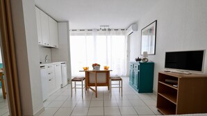 A modern kitchen with white cabinets, a dining table with two chairs, a teal cabinet, and a TV on a wooden stand. - Studio Héliopolis D 51 - 4 people (AGDE)
