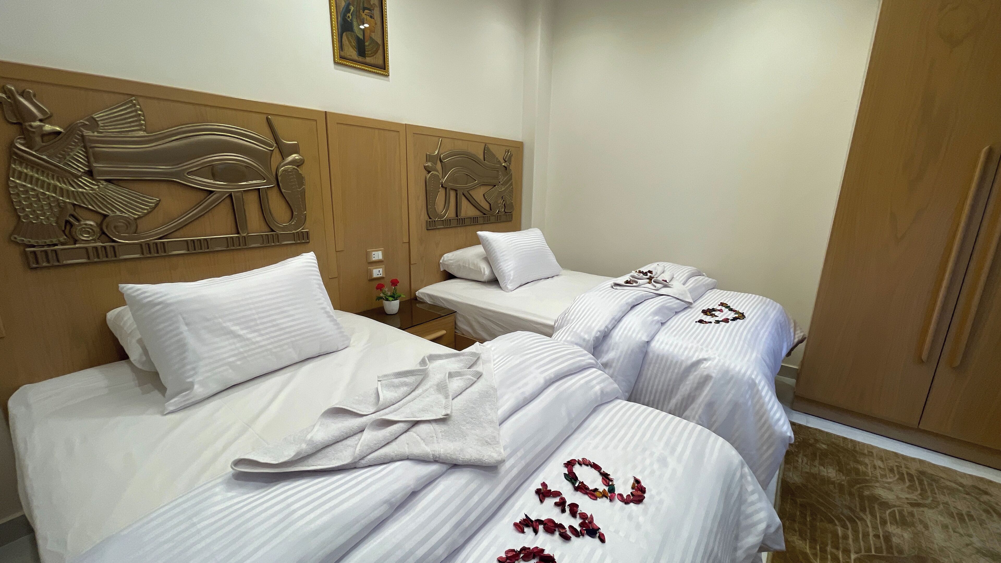 Een hotelkamer met twee bedden, elk met witte lakens en decoratieve kussens. Er is een houten hoofdeinde met decoratieve gravures, een kleine tafel met een bloemenvaas en een houten garderobe.