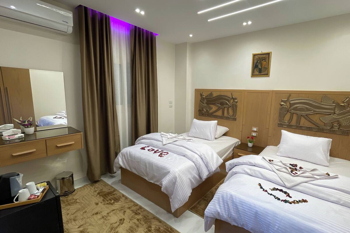A hotel room with two beds, a wooden headboard with carvings, a large mirror, a bedside table with a lamp, and a small potted plant.