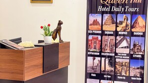 A hotel reception desk with a framed picture of an Egyptian figure, a cat statue, and a potted plant. A large poster advertises daily tours at the hotel, featuring landmarks like the Giza Pyramids and the Egyptian Museum. - Queen Inn Pyramids View (Giza)