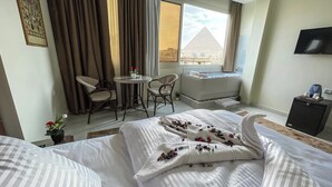 A hotel room with a bed, a small table with chairs, a TV, and a window with a view of a pyramid. - Queen Inn Pyramids View (Giza)