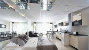 Quarto de hotel moderno com cama grande, cozinha compacta, área de jantar e sala de estar com uma janela ampla.