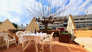 Une terrasse extérieure avec une table dressée pour deux personnes, entourée de chaises en plastique blanches, d’un grand bac à fleurs rempli de verdure et d’un arbre qui offre de l’ombre.