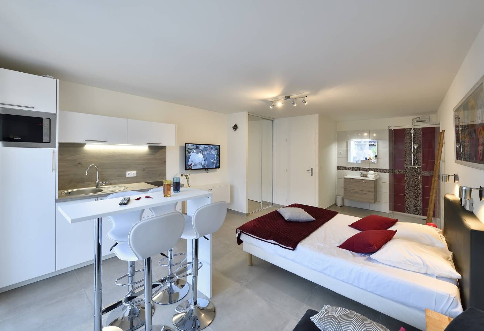A modern hotel room with a kitchenette, a bed with red and white bedding, a TV mounted on the wall, and a shower area.