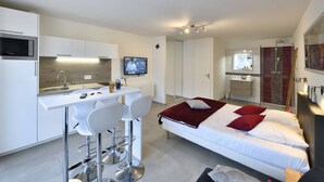 A modern hotel room with a kitchenette, a bed with red and white bedding, a TV mounted on the wall, and a shower area. - GENTLEMAN - Studio Héliopolis D 84 - Luxury - 2 people (AGDE)