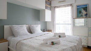 Ein ordentlich bezogenes Bett mit weißer Bettwäsche und aufgerollten Handtüchern, ein Nachttisch aus Holz und ein Fenster mit transparenten Vorhängen.