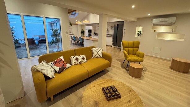 Ein modernes Wohnzimmer mit einer Couch, einem gelben Sessel und einem Holztisch. Im Hintergrund ist eine Essecke und eine Küche erkennbar.