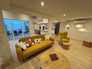 Un salon moderne avec un canapé, un fauteuil jaune et une table basse en bois. On aperçoit à l’arrière-plan un coin repas et une cuisine.