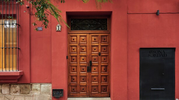 A red building with a large wooden double door, a black door to the right, and a window with a metal grill on the left.