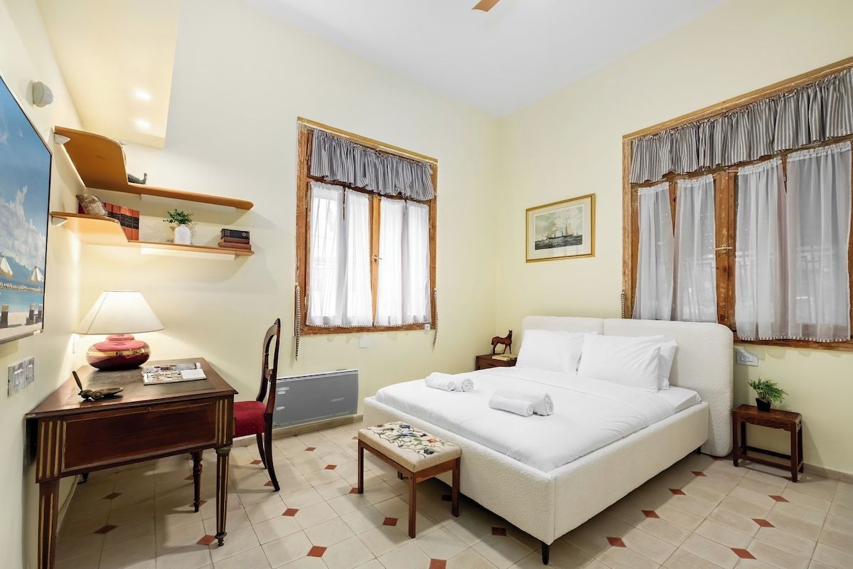 A hotel room with a bed, a desk, a chair, a small table, and two windows with curtains.