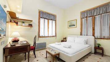 A hotel room with a bed, a desk, a chair, a small table, and two windows with curtains.