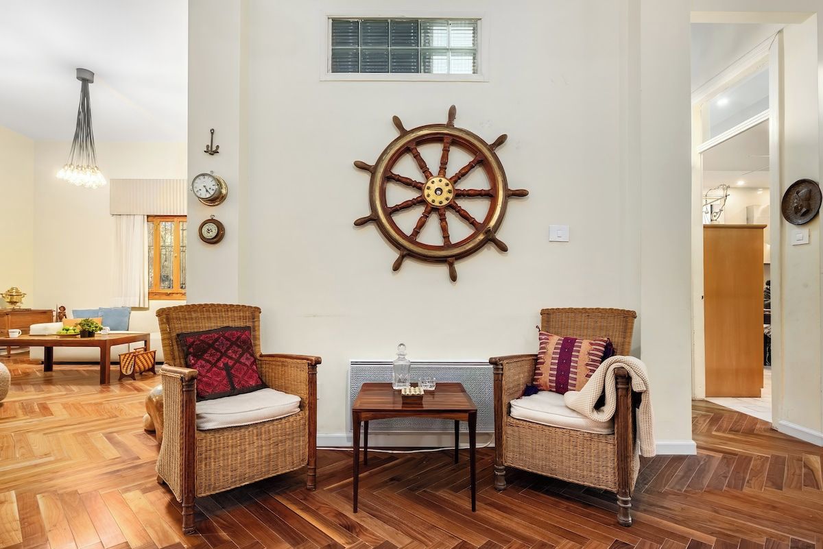 A room with a wooden floor, two wicker chairs with cushions, a wooden table, a wall-mounted ship's wheel, a window, and a hanging light fixture.