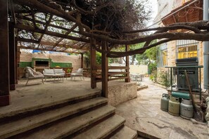 A covered outdoor seating area with steps leading up to it, surrounded by trees and a building with a green-tiled wall.