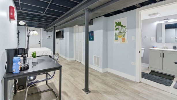 Una habitación de hotel moderna con cama, escritorio con lámpara, planta pequeña y baño con espejo y lavamanos.