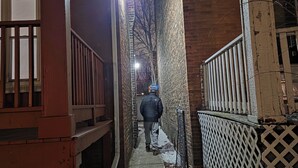 Un estrecho callejón flanqueado por edificios de ladrillo, una cerca de madera, y una persona alejándose de la cámara.