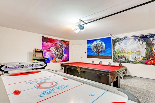Una mesa de billar con superficie de fieltro rojo, una mesa de fútbol de mesa y una gran mesa de hockey sobre hielo en una habitación con obras de arte en las paredes.