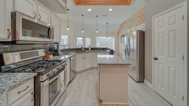 Una cocina moderna con armarios blancos, electrodomésticos de acero inoxidable, encimeras de granito y techo de madera.