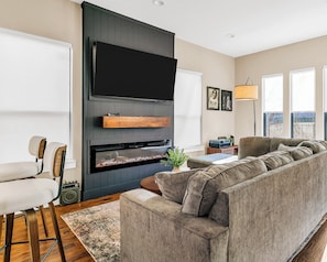 A modern living room with a large flat-screen TV mounted on a dark wall, a built-in fireplace, a grey sectional sofa, and a small side table with a lamp. - Dallas escape - Rooftop + Hot tub - Vintage Arcade (Dallas)