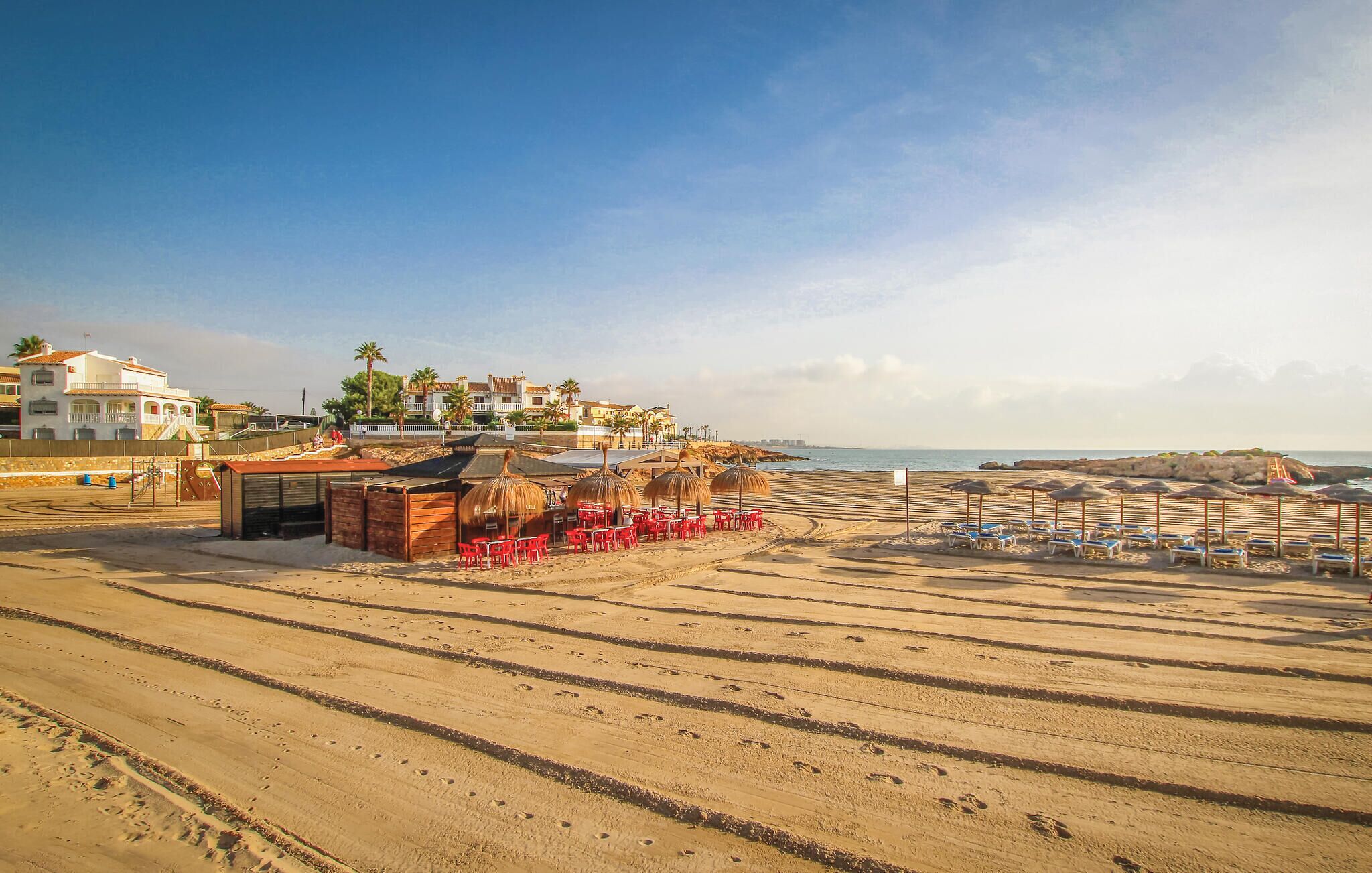 Une plage avec des chaises et des parasols rouges, un immeuble doté de balcons et un ciel dégagé.