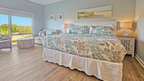 A bedroom with a large bed, a ceiling fan, a view to the outside, and a nightstand with a lamp. - Luau 6312 Cozy Studio Retreat | Pool, Tram, Beach | Skis & Seas (Miramar Beach)