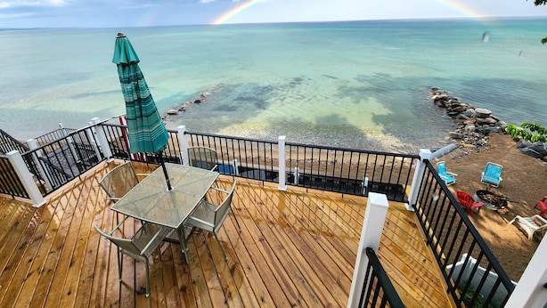 A wooden deck with a table and chairs, overlooking a beach with clear water and a rainbow. - 🌅 Sunset+Kayak | Private Beach House | M22 ➠ 7633 (Traverse City)