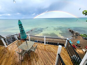 Una terraza de madera con una mesa y sillas, con vistas a una playa de aguas cristalinas y un arcoíris.