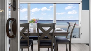 A dining area with a table set for four, a vase with flowers, and a view of the sea through large windows. - 🌅 Sunset+Kayak | Private Beach House | M22 ➠ 7633 (Traverse City)