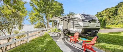 A recreational vehicle (RV) parked on a grassy area with a white fence, red Adirondack chairs, and a picnic table.
