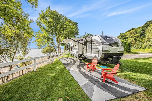 A recreational vehicle (RV) parked on a grassy area with a white fence, red Adirondack chairs, and a picnic table. - 🌅 Stars+Sunsets | Luxury Camper | Private Beach M22 ➠ 7633 (Traverse City)