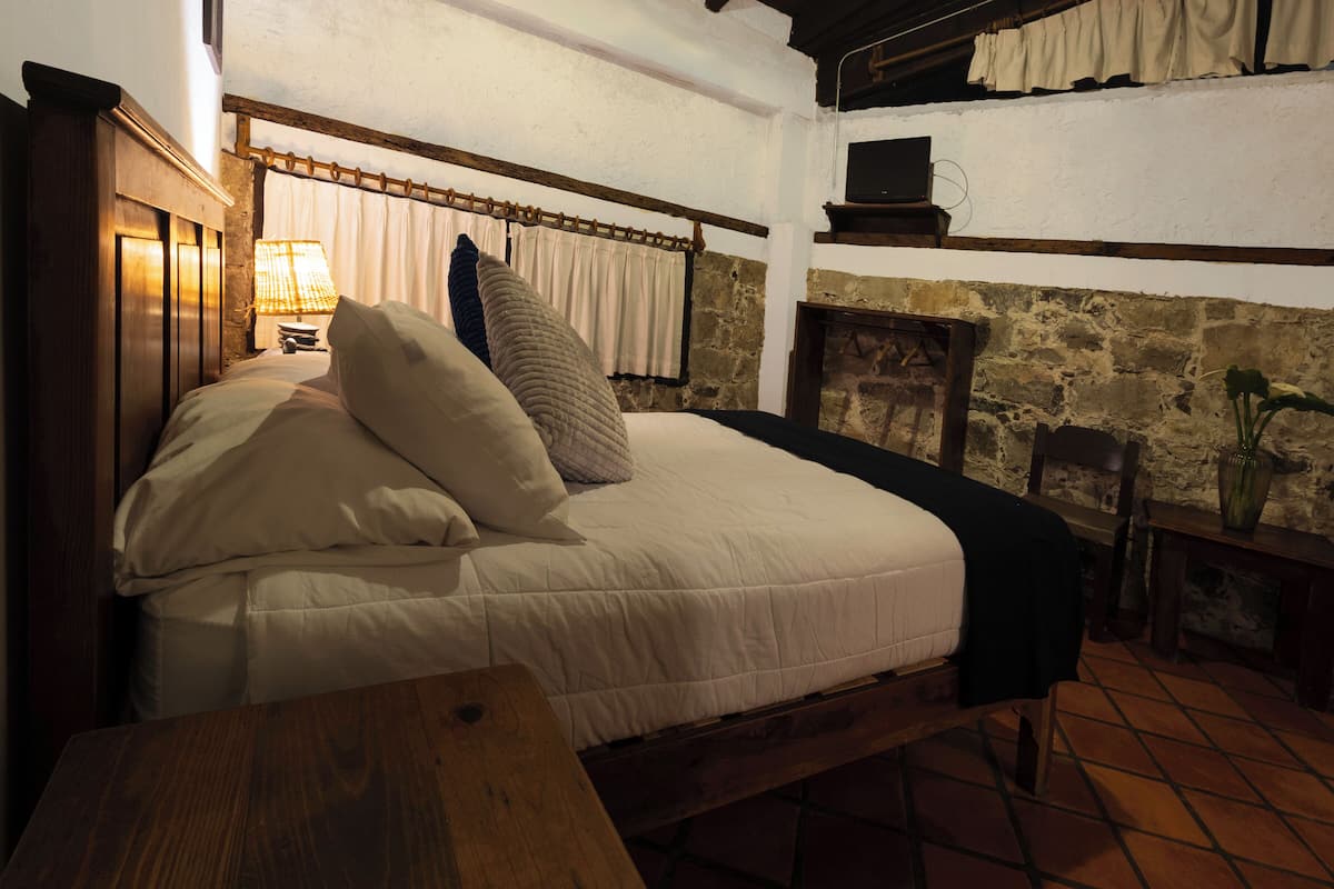 A cozy room with a bed, a wooden table, a lamp, and a stone wall.