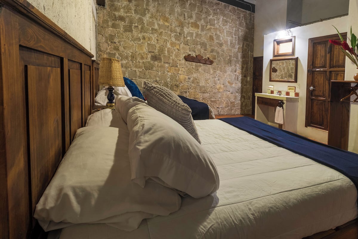 A neatly made bed with white linens and multiple pillows, a wooden headboard, and a stone wall in the background.