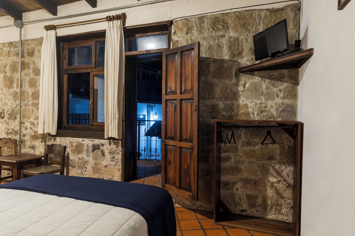 A rustic room with a bed, wooden furniture, a television mounted on a shelf, and a stone wall.