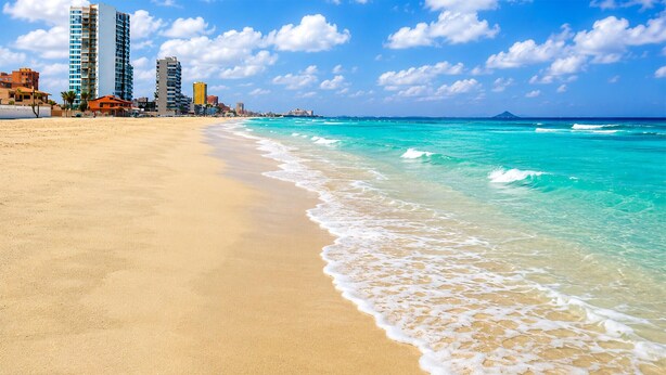 Una playa con aguas turquesas cristalinas, una orilla arenosa y una hilera de edificios al fondo.