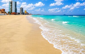 Ein Strand mit klarem türkisblauem Wasser, einem sandigen Ufer und einer Reihe von Gebäuden im Hintergrund.