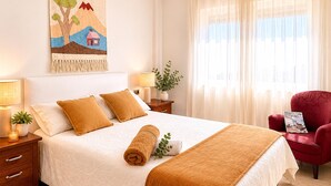 Una cama bien tendida con una manta blanca y marrón, dos almohadas, una toalla enrollada, una planta pequeña y un sillón rojo.
