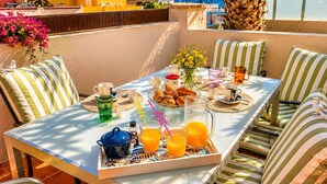 Una terraza iluminada por el sol con una mesa lista para el desayuno, cojines a rayas y vistas al mar.