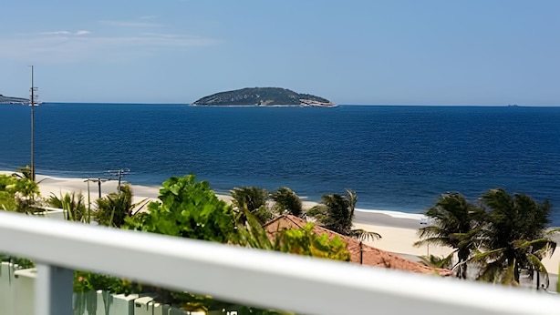 Blick auf einen Strand mit einer kleinen Insel im Hintergrund, einem klaren Himmel und etwas Grün im Vordergrund.