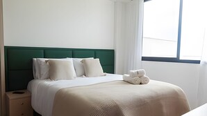 Un lit bien fait, avec une tête de lit verte, une literie blanche et une table de chevet sur laquelle se trouve une lampe.