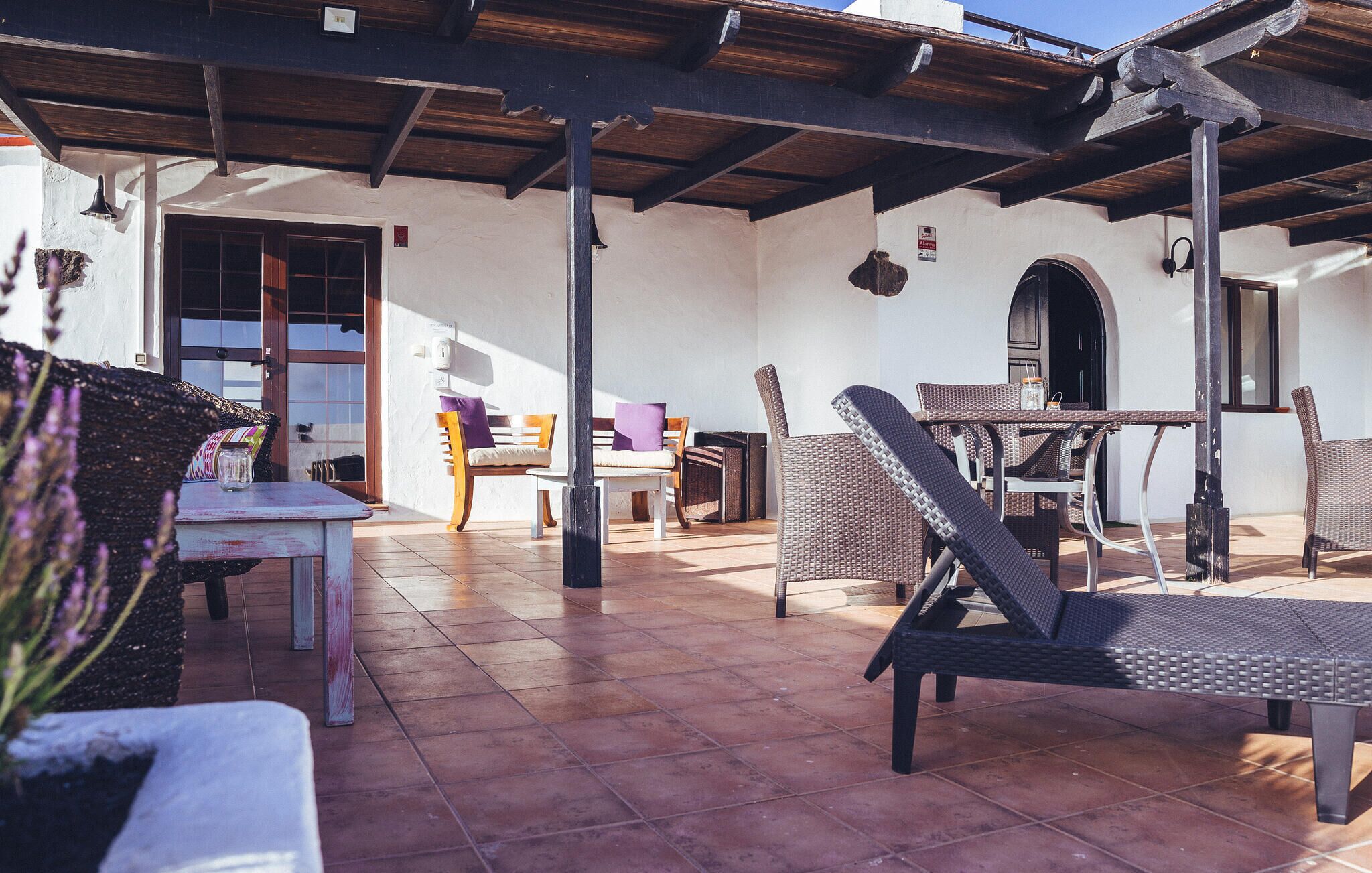 Un espace extérieur aménagé en terrasse, avec des meubles en osier, une table et un banc. Une porte donne accès à l’intérieur.