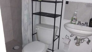 Ein kleines Badezimmer mit Toilette in Weiß, einem grünen Mülleimer, einem schwarzen Metallregal, einem weißen Waschbecken und einer Duschkabine.
