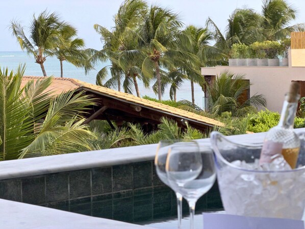 A table with a bottle of champagne and a glass, overlooking a beach with palm trees and a wooden deck.