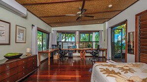 Une chambre spacieuse avec un grand lit, un bureau avec deux chaises et un ordinateur. Des fenêtres offrent une vue sur la verdure environnante.