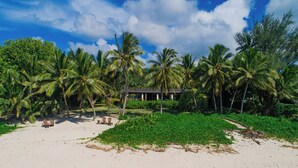 Une propriété située en bord de mer, agrémentée de palmiers, d’une plage de sable et d’un bâtiment doté d’un toit foncé.