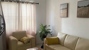 En stue med en beige sofa, en mindre lænestol, et glasbord, en potteplante og to billeder i rammer på væggen.