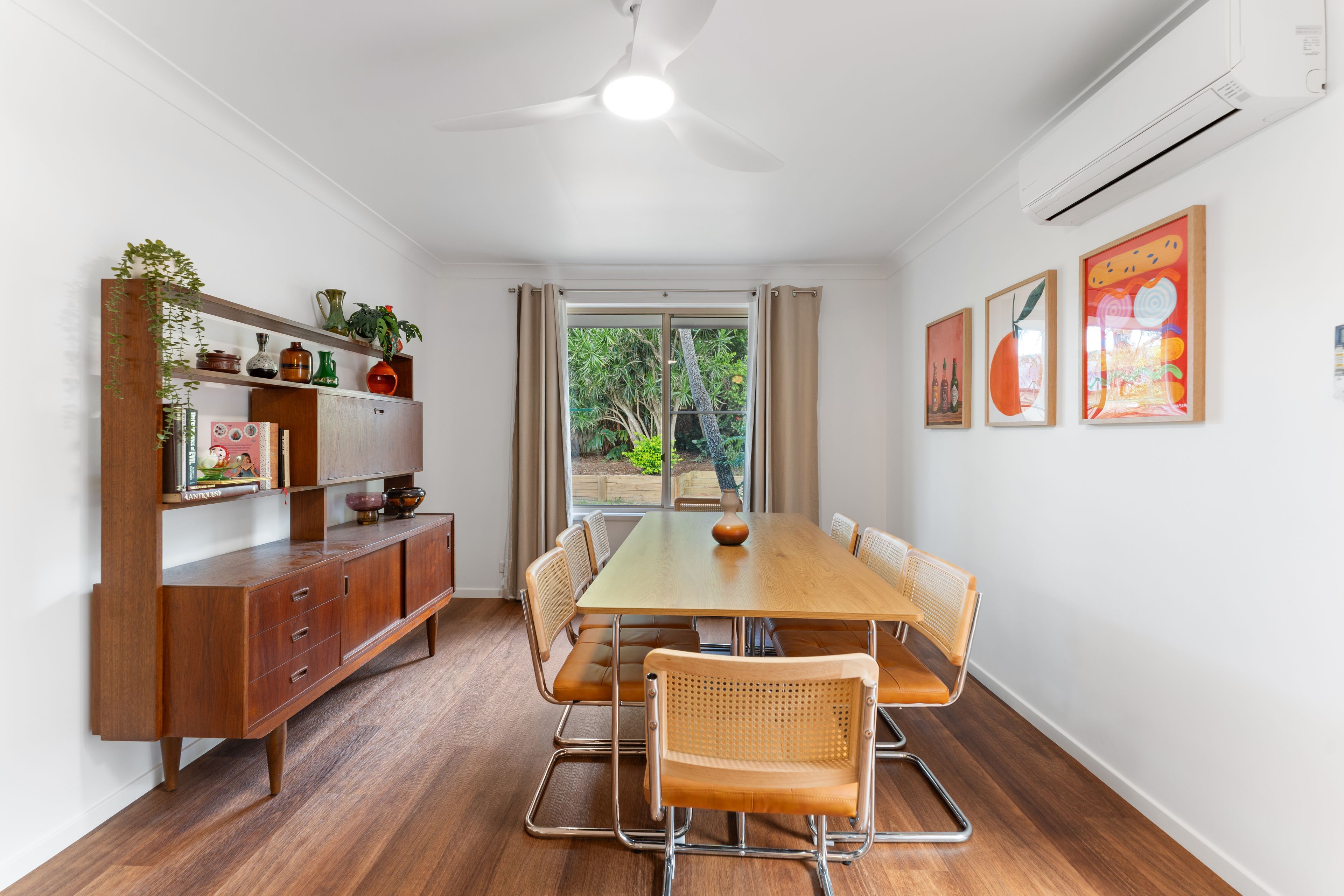 A dining area with a wooden table, six chairs, a ceiling fan, and a view of greenery outside.