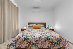 Ein Schlafzimmer mit geblümter Bettdecke, einem Holz-Kopfteil, einem Nachttisch mit Lampe und einer Wand-Klimaanlage.