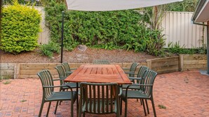 Un patio con tavolo e sedie in legno, circondato da un pavimento in mattoni e un giardino con diverse piante e una recinzione in legno.